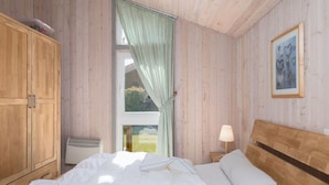 A bedroom with wooden walls, a large window with curtains, a bed with white bedding, a bedside table with a lamp, and a framed picture on the wall. - Ferienhaus mit Sauna im Ferienpark Mirow (Mirow)
