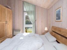 A bedroom with wooden walls, a large window with curtains, a bed with white bedding, a bedside table with a lamp, and a framed picture on the wall.