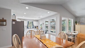 Un coin repas avec une table et des chaises en bois, une cuisine à l’arrière-plan et de grandes fenêtres.