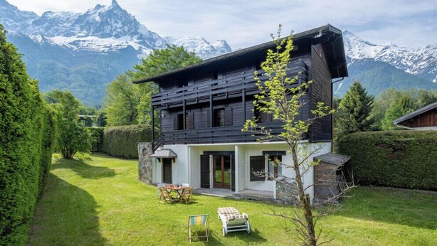Une maison moderne à deux étages, dotée de fondations en pierre, entourée d’une pelouse et de haies bien entretenues, et qui se dresse sur un fond de montagnes enneigées.