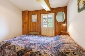 Une chambre avec un mur lambrissé en bois, un lit recouvert d’une couvre-lit à motifs, une porte avec une fenêtre, un miroir rond et une lampe de chevet.