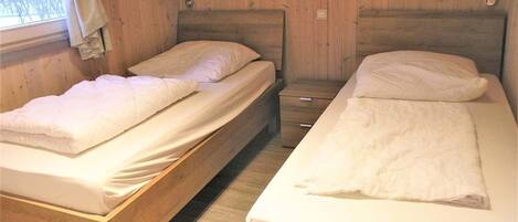 Two single beds with wooden frames and white bedding in a room with wooden walls and a window with curtains.