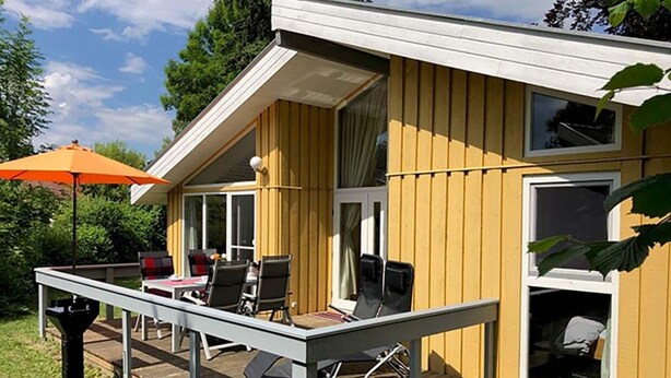A yellow wooden house with a deck, outdoor furniture, and a large window. - Ferienhaus mit Sauna im Ferienpark Mirow (Mirow)