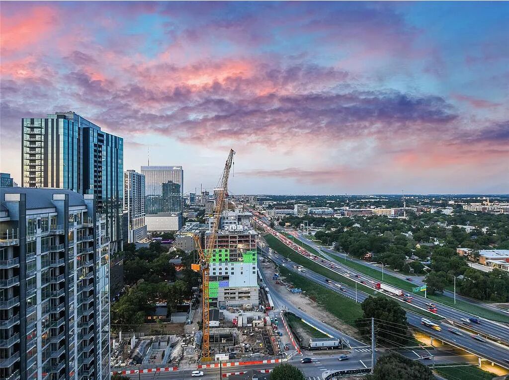 A high-rise building with a construction crane, surrounded by other tall buildings, under a colorful sky.