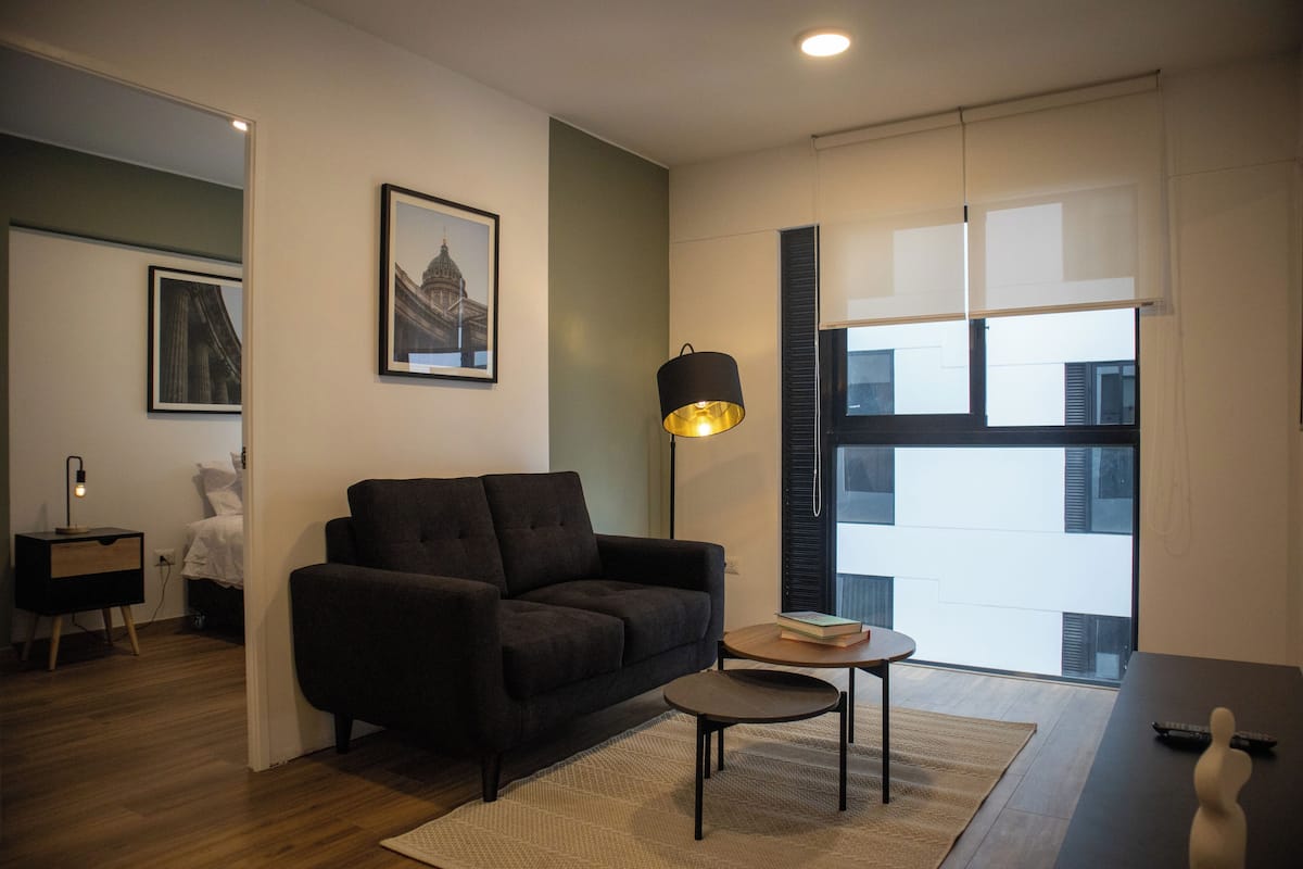 Una sala moderna con un sofá oscuro, una mesita redonda pequeña y una lámpara de pie. Hay un cuadro enmarcado en la pared y una ventana con persianas.