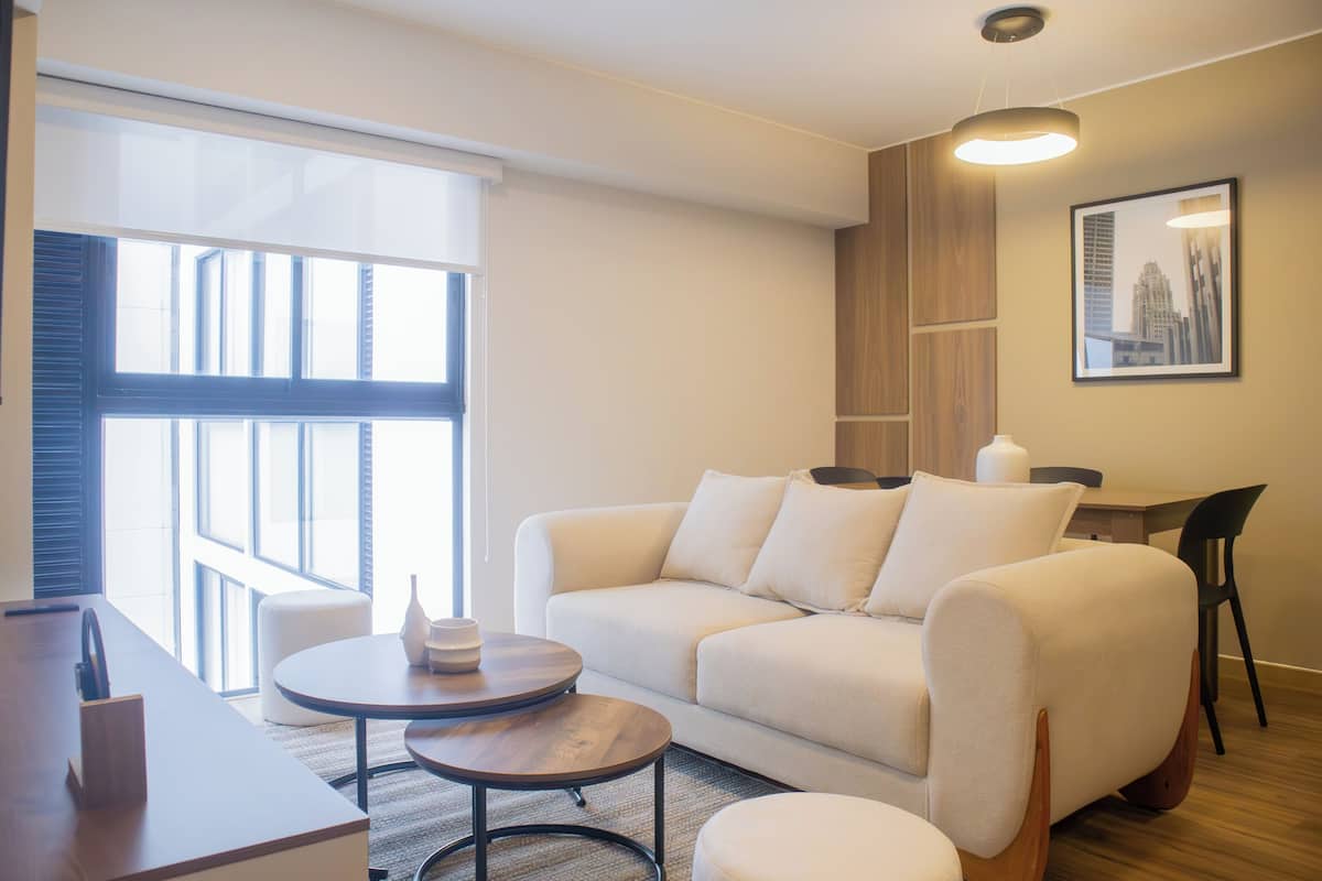 Una sala de estar moderna con sofá, mesa de centro y reposapiés, una mesa de comedor con sillas y un amplio ventanal con persianas.