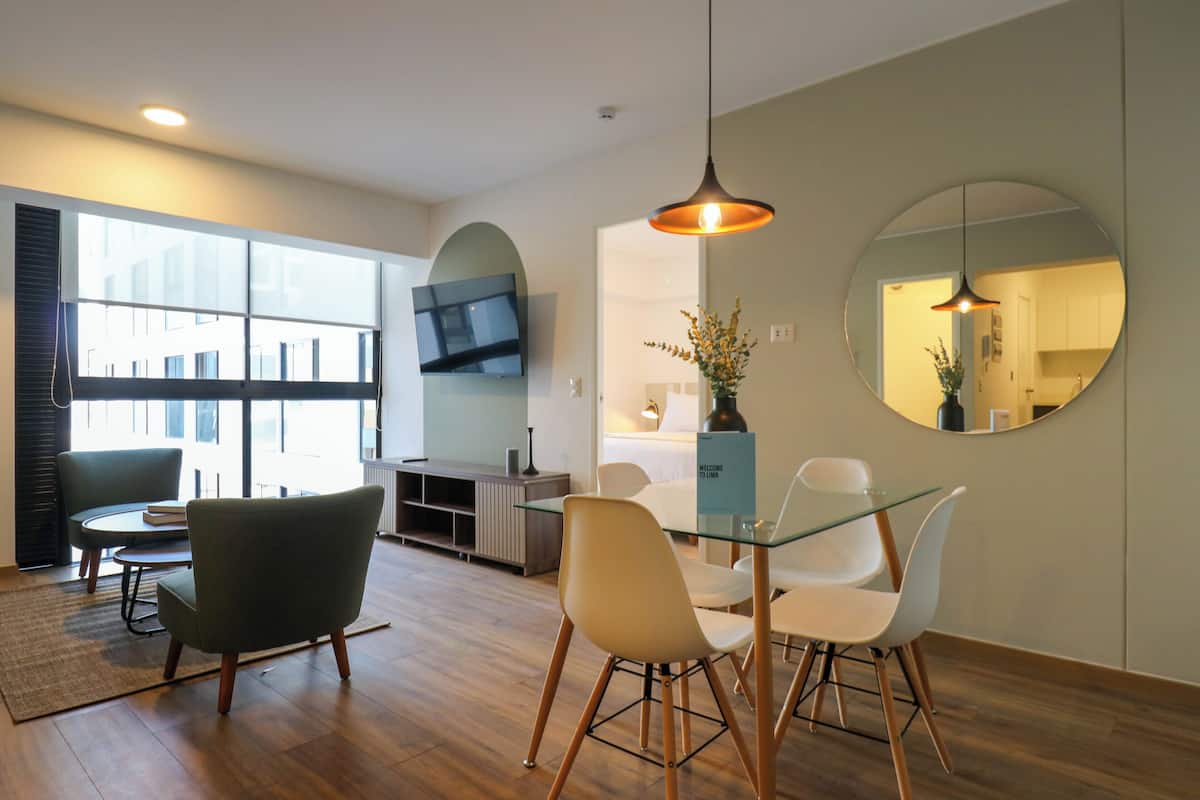 Una sala de estar moderna con una mesa de comedor de vidrio, dos sillas, un sofá y un televisor montado en la pared.