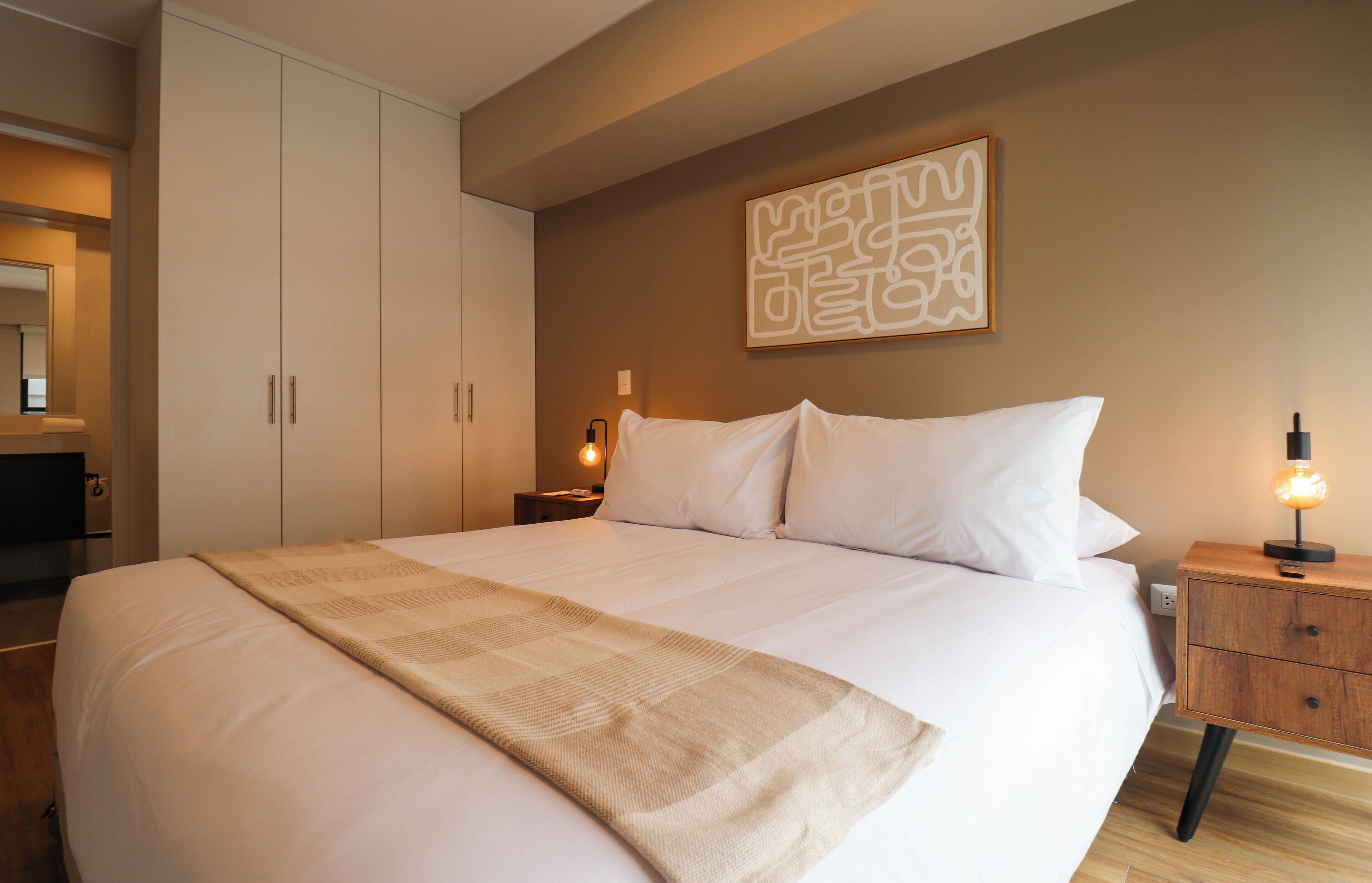 Una cama bien tendida con una colcha blanca y un plaid color beige, una mesita de noche con una lámpara, y una obra de arte abstracto colgada en la pared.