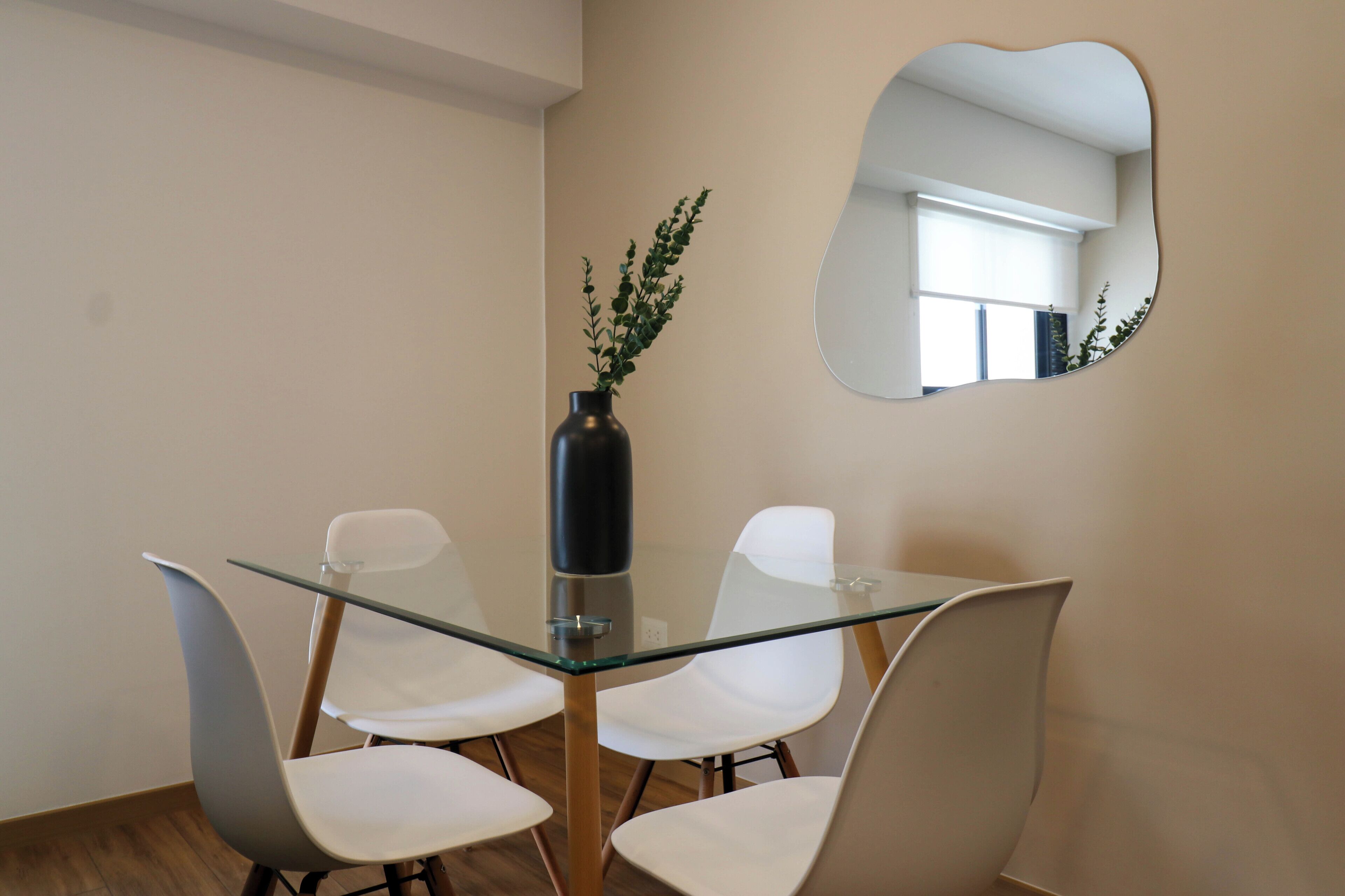 A modern dining area with a glass table, white chairs, a vase with green plants, and a unique mirror on the wall.