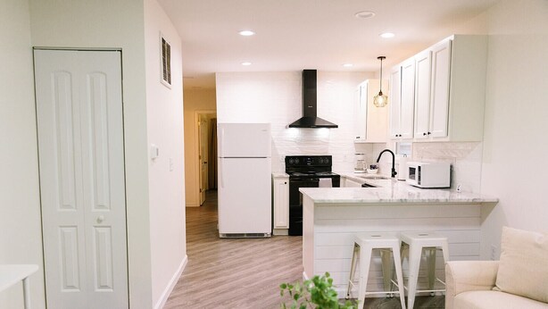 A modern kitchen with white cabinets, a black stove, and a white refrigerator. There is a small island with stools and a microwave. - Goldenrod - Studio in Downtown Lexington (Lexington)