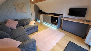 A modern loft-style living space with a wooden ceiling, a gray sofa, a kitchen area with a built-in oven, and a flat-screen TV. - Galloway Steading Luxury Glamping Pods (Newton Stewart)