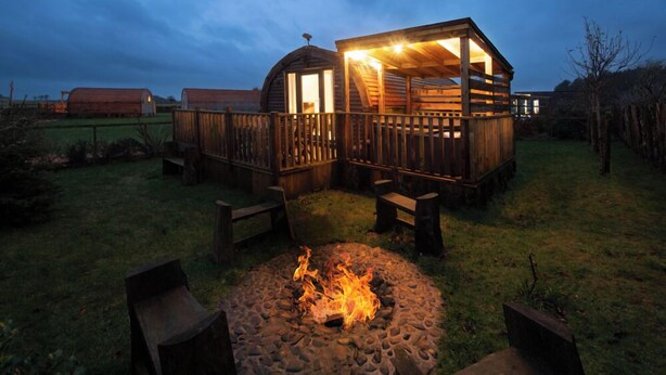 A rustic wooden cabin with a fire pit in the foreground, surrounded by a wooden deck and benches. - Galloway Steading Luxury Glamping Pods (Newton Stewart)