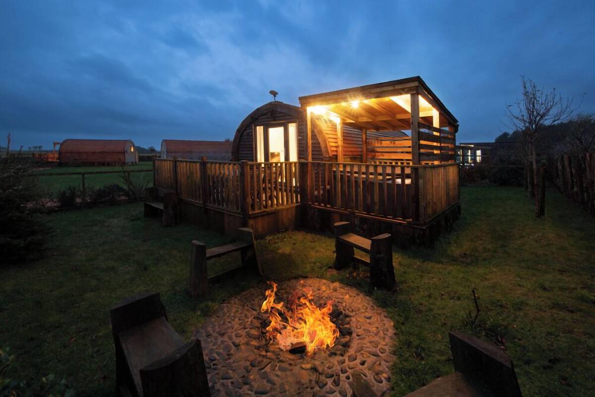 A rustic wooden cabin with a fire pit in the foreground, surrounded by a wooden deck and benches.