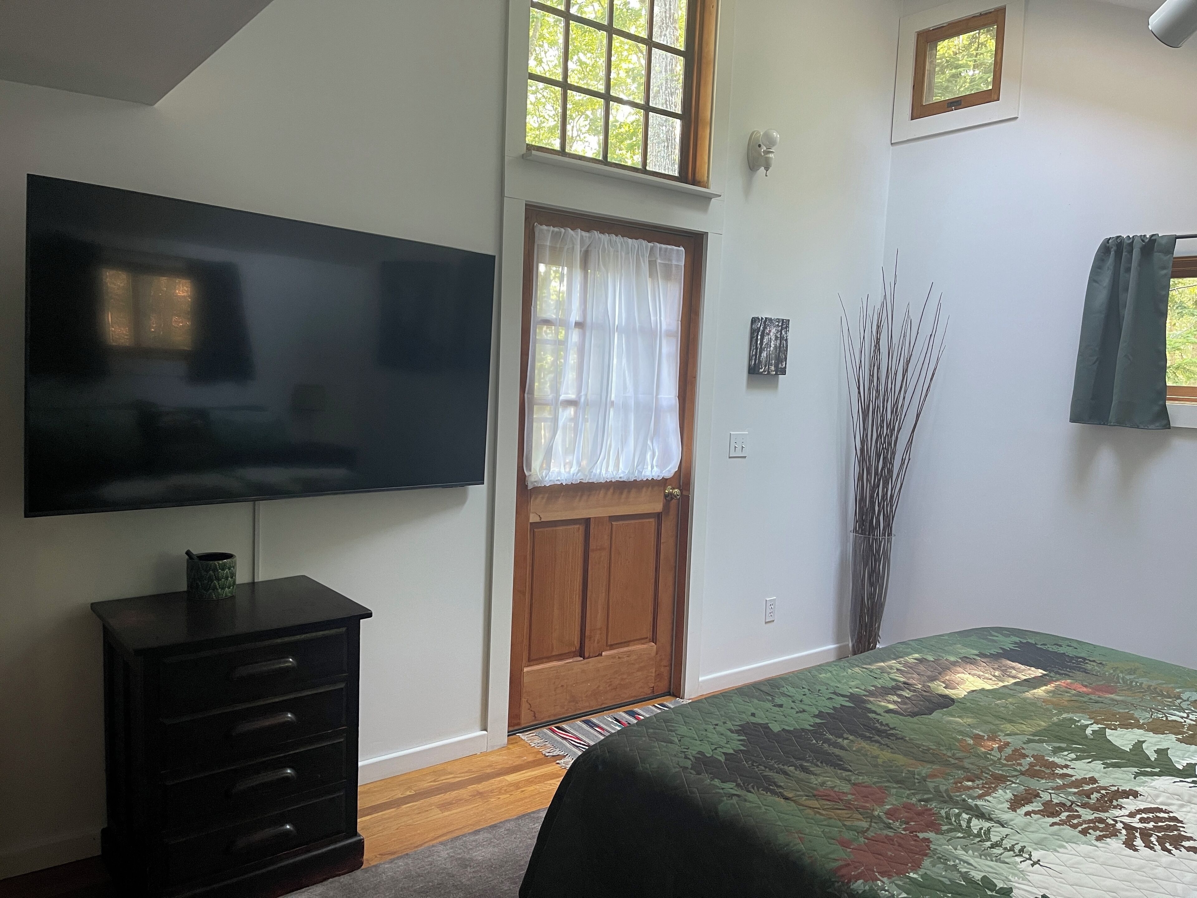 A bedroom with a large flat-screen TV, a wooden door, a window with sheer curtains, a bed with a floral bedspread, and a tall vase of branches.