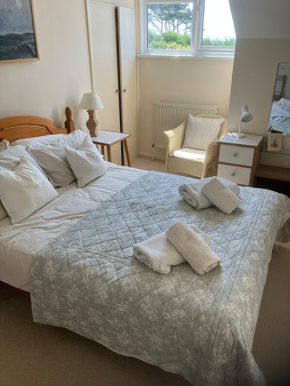 A neatly made bed with a floral quilt, a chair, a bedside table with a lamp, and a window with a view of greenery.