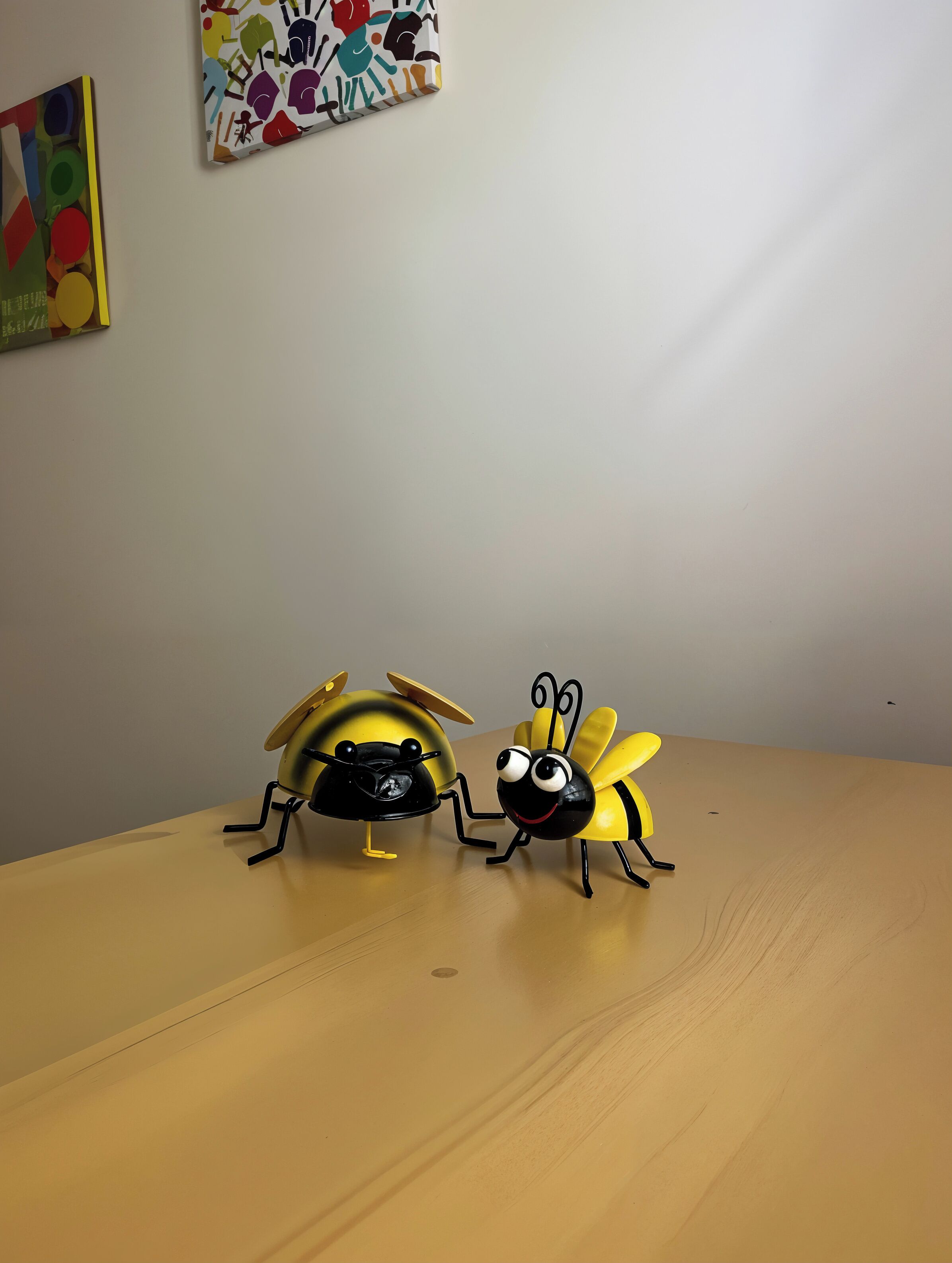 Two toy bees on a wooden table with a plain wall and a colorful painting in the background.