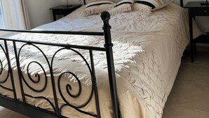 Cama bem arrumada com estrutura de metal preto e colcha bege, mesa de cabeceira com abajur, e quadro na parede com casas coloridas.