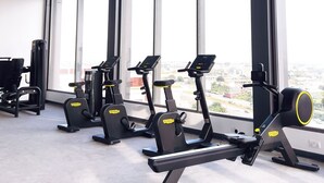 Academia com equipamentos de ginástica, janelas amplas e vista para a cidade.