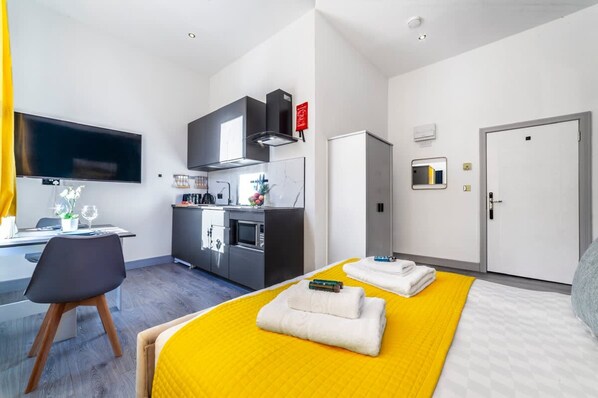 A modern hotel room with a bed, a desk, a kitchenette, and a TV.