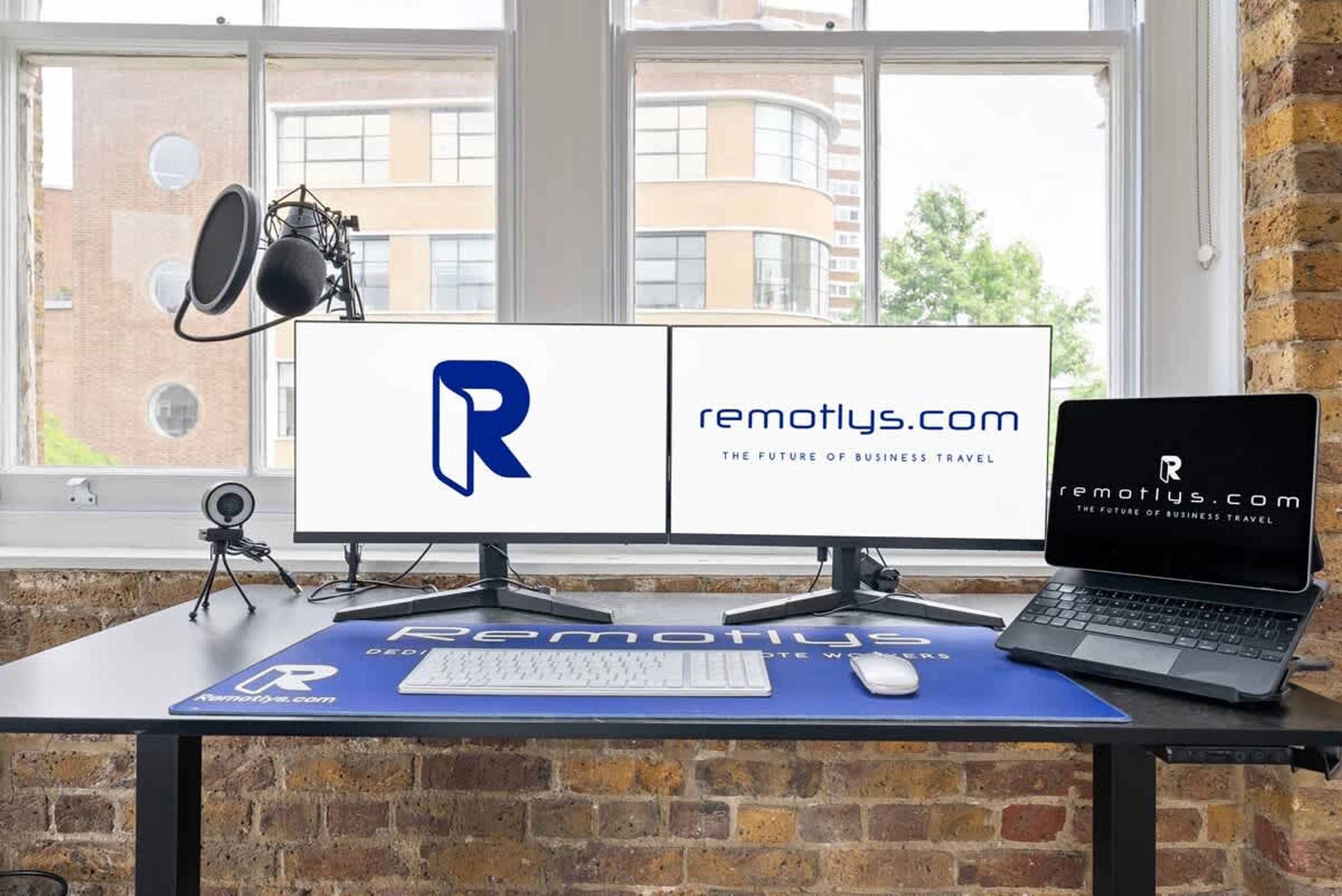 Un espacio de trabajo con un monitor de computadora mostrando el logotipo de Remotely.com, una computadora portátil, un micrófono y una cámara web sobre un escritorio.