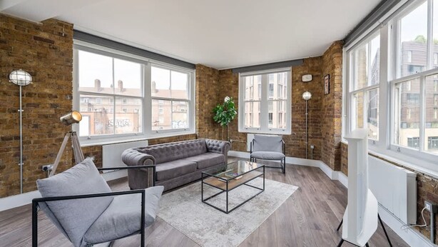 A modern living room with a sectional sofa, a glass coffee table, and a white armchair. Large windows provide natural light and views of the city. - Spacious Bright Oasis in Old Street (London)