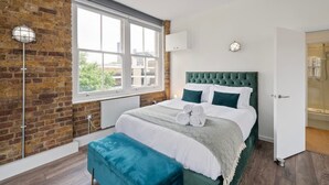 A bedroom with a brick wall, a large window, a green upholstered headboard, and a bench. - Spacious Bright Oasis in Old Street (London)