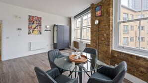 A modern living room with a glass dining table, grey chairs, a brick wall, a radiator, and a large window with a city view. - Spacious Bright Oasis in Old Street (London)