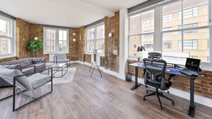 A modern office space with a brick wall, large windows, a grey sofa, a desk with a computer, and a standing lamp. - Spacious Bright Oasis in Old Street (London)
