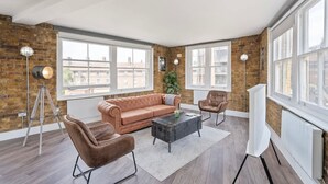 Una sala de estar moderna con un sofá de cuero, una mesa de centro y dos sillones. Hay ventanas grandes y una pared de ladrillo.
