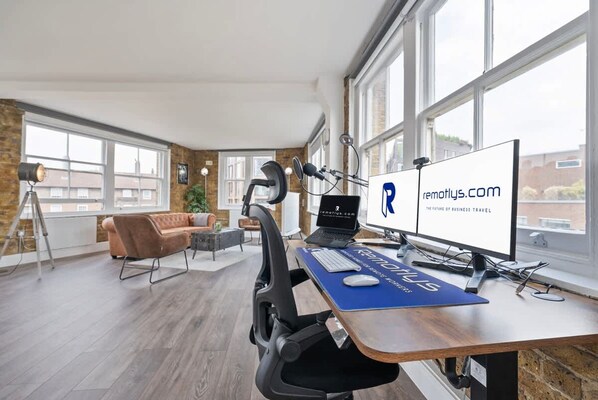 A modern office with a large desk, computer, and headphones. There is a couch and a plant in the background. - Large 1 Bedroom Studio in Old St (London)