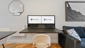 Una habitación moderna con un escritorio, dos monitores, una silla, un sofá y una obra de arte enmarcada en la pared.