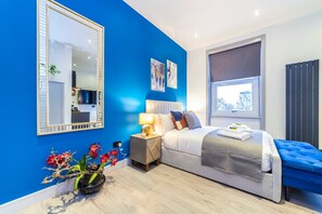 A modern bedroom with a large mirror, a bed with a blue accent, a bedside table with a lamp, and a window with a view. - Studio 4 - Cosy Studio in the Heart of Earls Court (London)