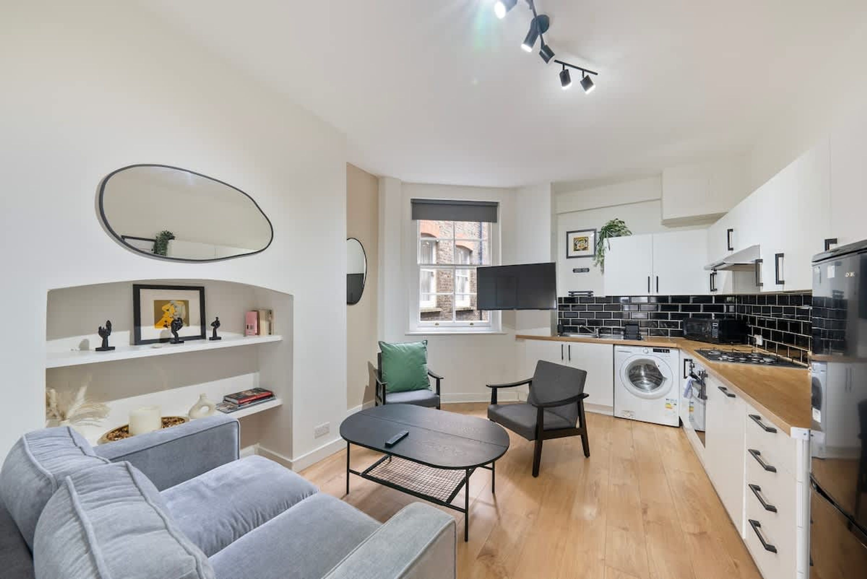 A compact living space with a sofa, a small table, a washing machine, a kitchen area, and a large mirror.