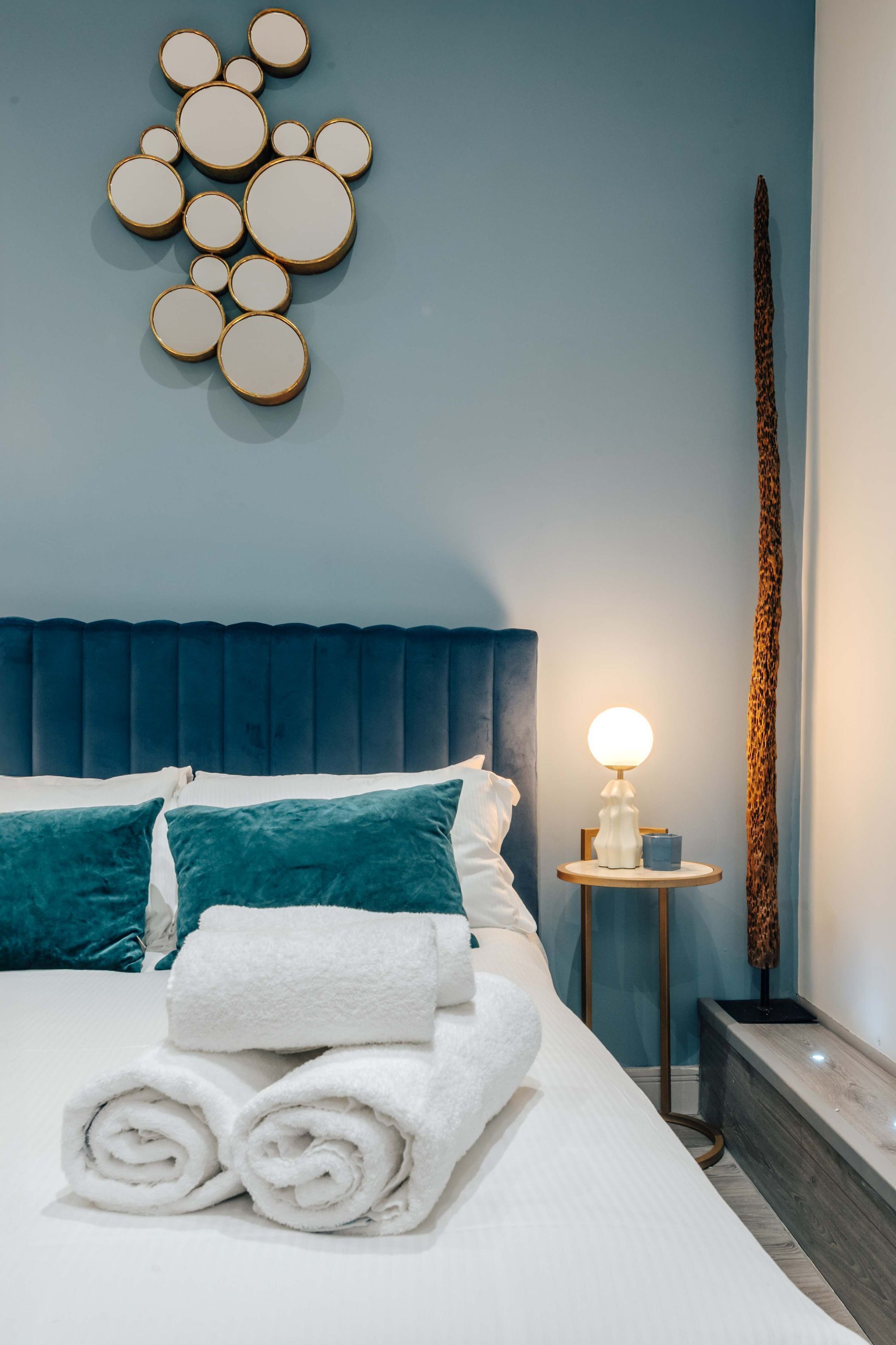 A neatly made bed with rolled white towels, a blue headboard, teal pillows, and a wall-mounted circular light fixture.