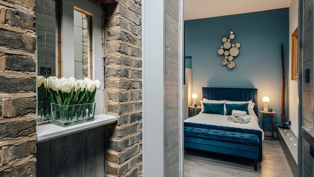 A bedroom with a bed, bedside tables, a wall-mounted light fixture, and a window with a flower arrangement. - Studio 1 - Charming Studio in Earls Court (London)