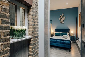 Un dormitorio con una cama, mesitas de noche, una lámpara de pared y una ventana con un arreglo floral.