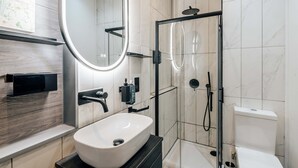 A modern bathroom with a large oval mirror, a white sink, a glass-enclosed shower, and a toilet. - Studio 1 - Charming Studio in Earls Court (London)