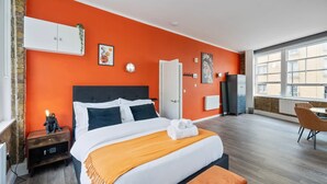 Ein modernes Schlafzimmer mit einem großen Bett, einem weißen Kleiderschrank, einem kleinen Nachttisch und einem Fenster mit Blick auf Gebäude.