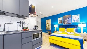 Une chambre d’hôtel moderne dotée d’un coin cuisine, d’un lit avec une literie jaune et bleue, et d’un mur d’accent bleu.
