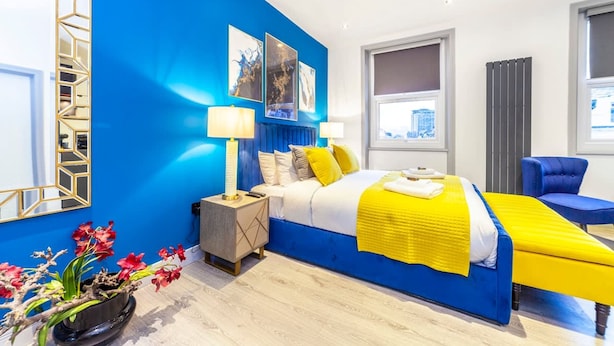 Une chambre d’hôtel moderne avec un mur d’accent bleu, un lit avec une literie jaune, une table de chevet avec des lampes et une vue sur la ville par la fenêtre.