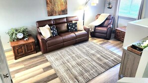 Ein Wohnzimmer mit einem braunen Ledersofa, einem Holztischchen mit einer Uhr, einem großen Gemälde, einer Stehlampe und einem Fenster mit Jalousien.