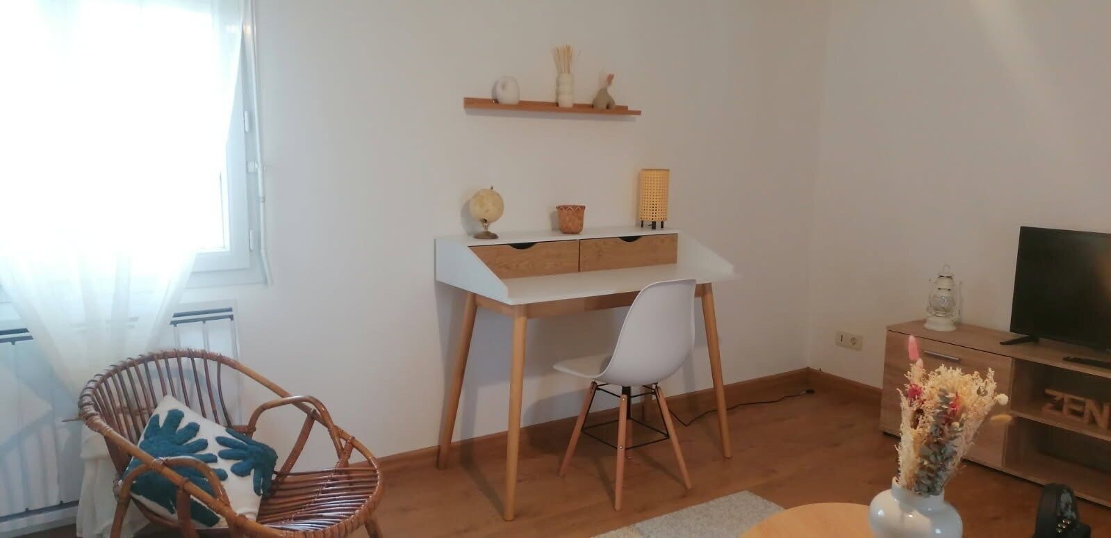 Een moderne woonkamer met een wit bureau, een rieten stoel, een witte stoel, een televisie en een plank met decoratieve objecten.