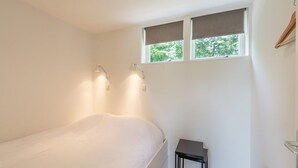 Quarto com cama, duas luminárias de cabeceira, uma mesinha, janela com persianas e radiador.