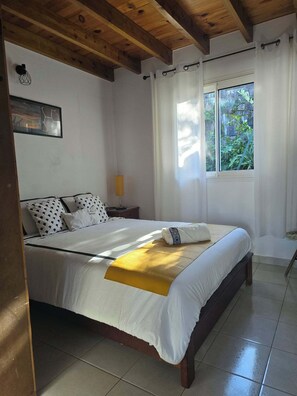 A bedroom with a bed, a nightstand, a lamp, a window with curtains, and a wooden ceiling.