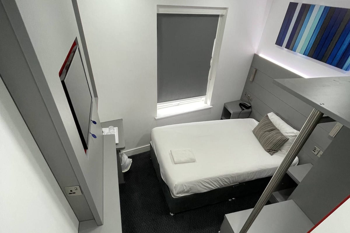 A compact hotel room with a bed, a small window, a bedside table, and a power outlet.