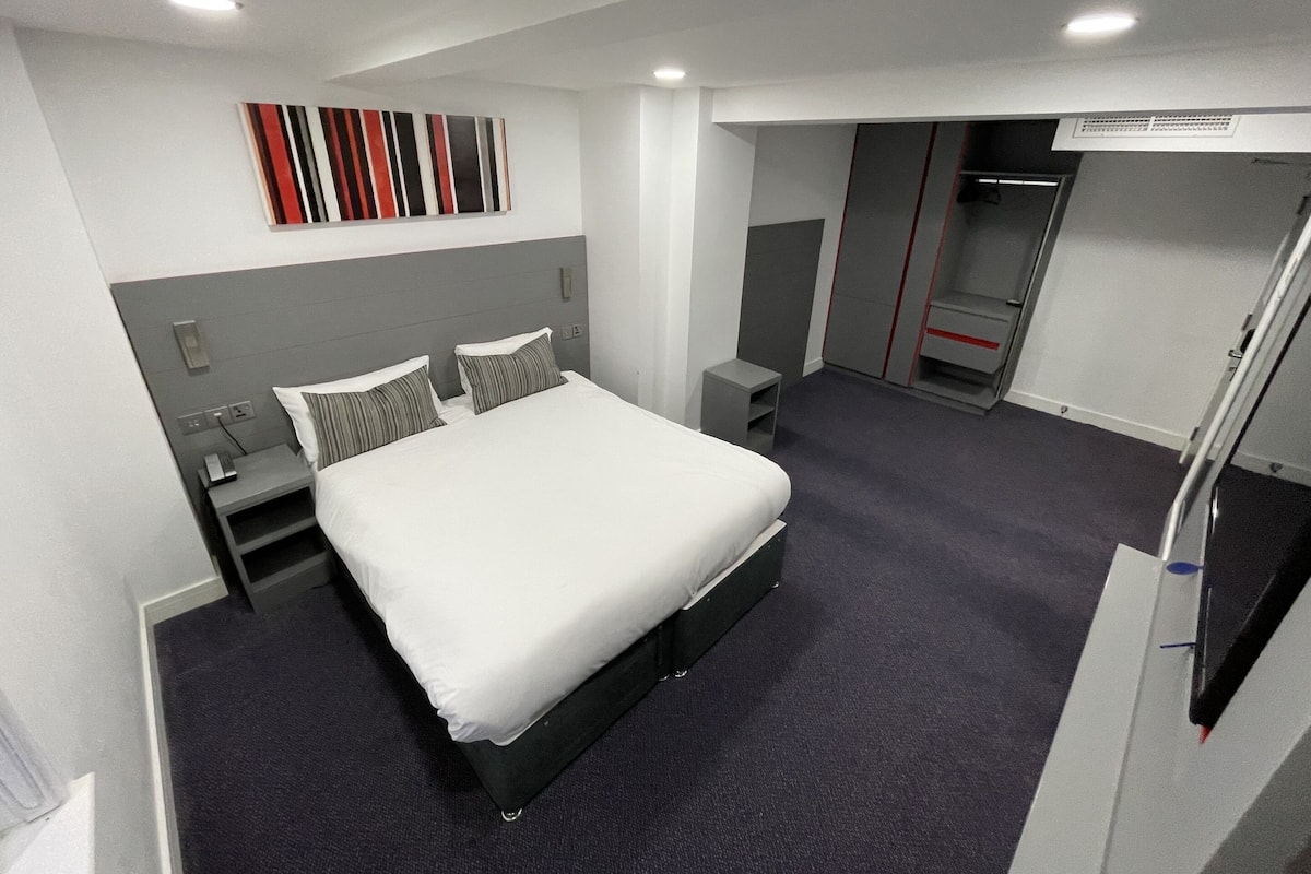 A modern hotel room with a large bed, a small bedside table, a nightstand, a wardrobe, and a mirror.