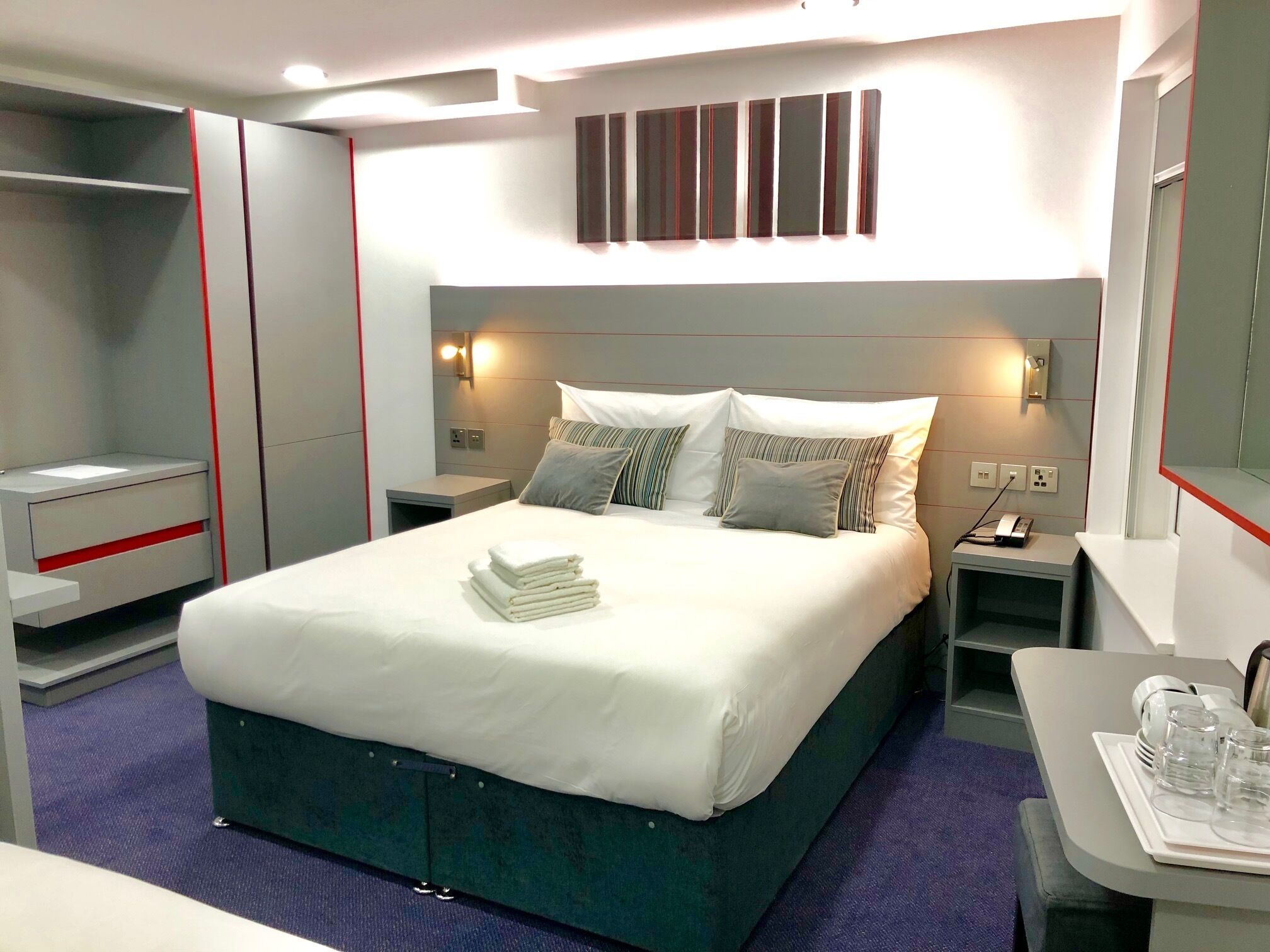 A modern hotel room with a neatly made bed, a bedside table with a lamp, a small desk with a chair, and a wall-mounted TV.