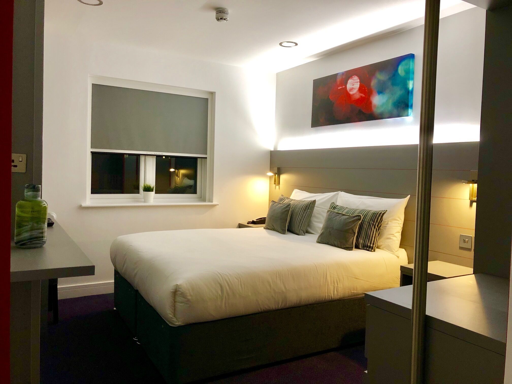 A modern hotel room with a large bed, bedside tables, a window with a view, and a painting on the wall.