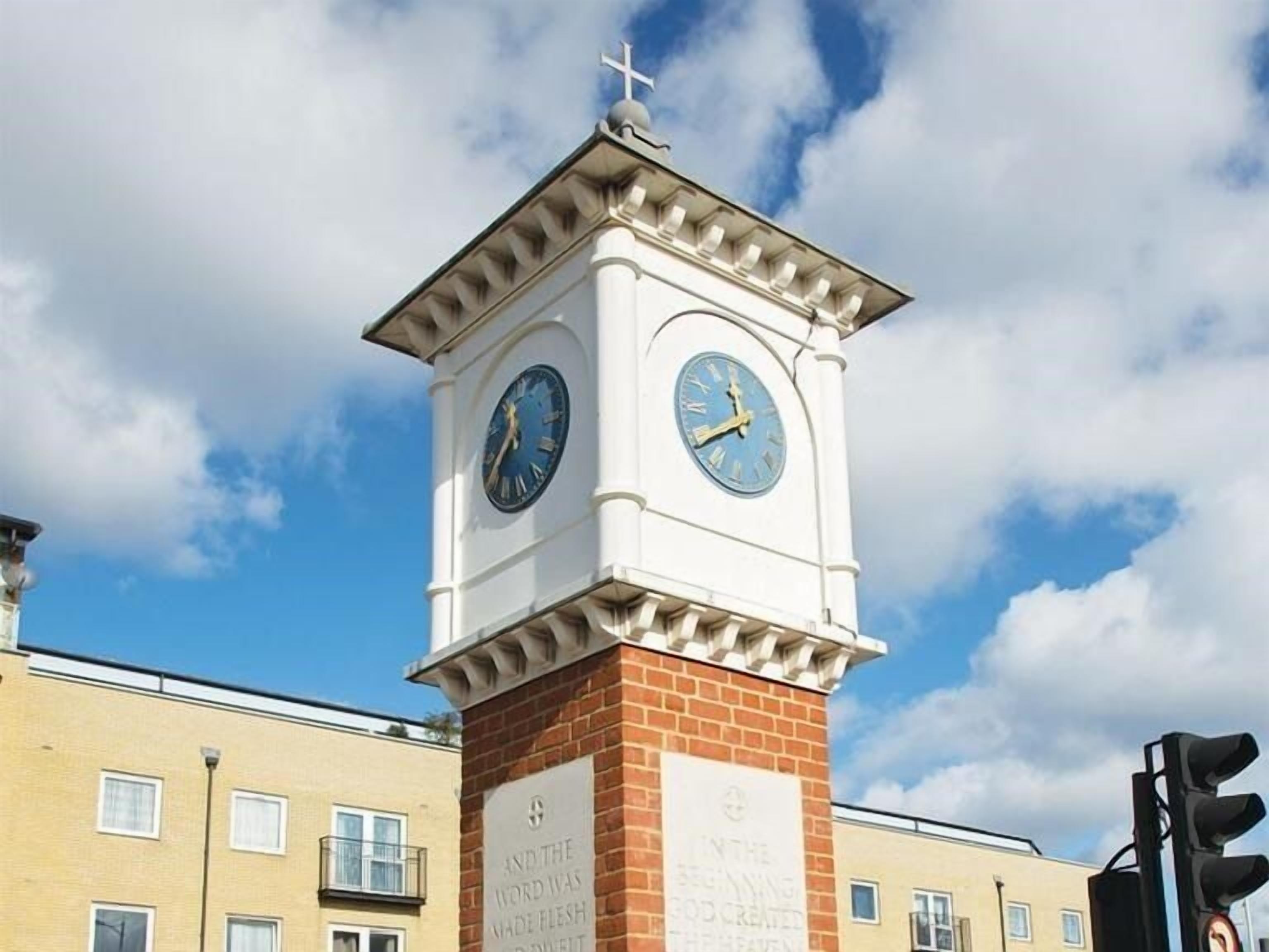 A clock tower with two faces, a cross on top, and a plaque with text.