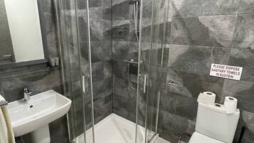 Une salle de bain moderne avec une douche en verre, un lavabo et des toilettes. Un panneau indique qu’il faut jeter les protections hygiéniques dans une poubelle.